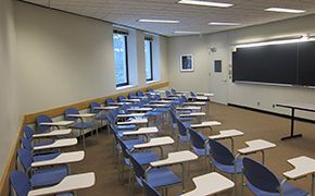 View from right side of classroom. A long table and three rows of chairs. A blackboard in front.