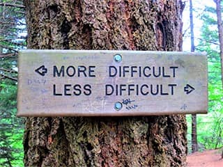 A tree with a block of wood fastened to it. Engraved on the block are the words: More Difficult, with an arrow pointing left, and Less Difficult, with an arrow pointing right.