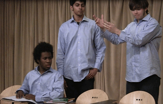 One student sits at a desk, while two others stand beside him, gesturing.