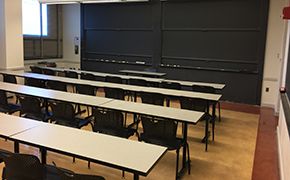 4 rows of white tables and black chairs facing a 2 sets of sliding chalkboards. A window on the left wall.