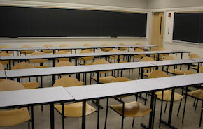 Four rows of narrow grey tables with wooden chairs. Blackboards at the back and side of the room