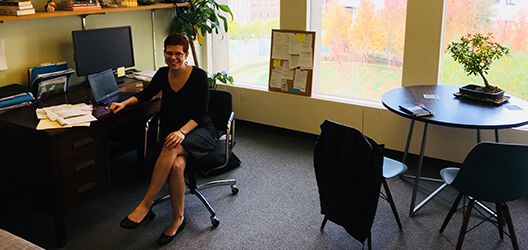 Woman sitting in a chair in front of a desk. Bookshelves on wall above desk. Windows on one wall. Fall foliage outside of widow.